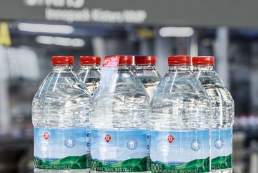 Das Bild zeigt mehrere Kunststoff-Wasserflaschen, die in einer Verpackungseinheit zusammengefasst sind, im Hintergrund befindet sich eine Verpackungsmaschine.