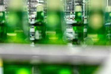 Das Bild zeigt unscharfe grüne Glasflaschen im Vordergrund, während im Hintergrund eine Abfüllanlage zu sehen ist.