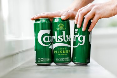 Das Bild zeigt drei Carlsberg-Dosen, die nebeneinander aufgestellt sind, während eine Hand die Dosen von oben berührt und leicht zusammendrückt.