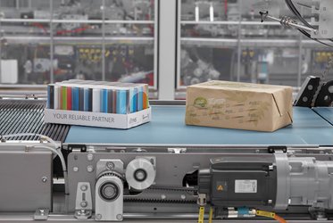 Das Bild zeigt zwei Verpackungen, eine aus Papier und eine in bunter Folie mit der Aufschrift "Your Reliable Partner", die auf einem Förderband in einer industriellen Produktionsanlage transportiert werden.
