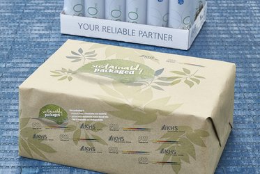 Das Bild zeigt eine umweltfreundliche Verpackung aus recycelbarem Papier mit der Aufschrift "sustainably packaged" auf einer Palette, die nachhaltige Verpackungslösungen von KHS bewirbt.