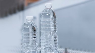 Das Bild zeigt zwei transparente Kunststoffflaschen in unterschiedlichen Größen mit weißen Verschlusskappen, die auf einem Förderband in einer Produktionsumgebung stehen und leichtes Flaschendesign repräsentieren.