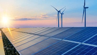 Das Bild zeigt ein Feld mit Solarpaneelen im Vordergrund und mehreren Windkraftanlagen im Hintergrund, während die Sonne am Horizont untergeht, was nachhaltige Energieerzeugung symbolisiert.