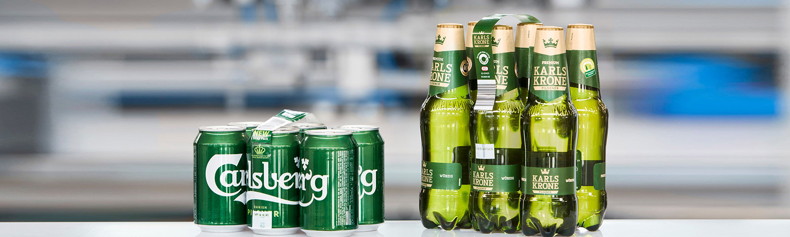 Das Bild zeigt zwei Verpackungsarten von Getränken: Links eine Carlsberg-Dose in einem Sechserpack mit minimaler Verpackung, und rechts eine Packung von sechs Karlskrone-Flaschen, die mit einem umweltfreundlichen, minimalistischen Tragegriff verbunden sind.