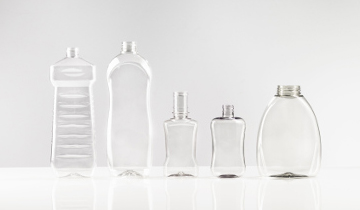Das Bild zeigt fünf transparente Kunststoffflaschen in unterschiedlichen Formen und Größen, die nebeneinander auf einer glatten, weißen Oberfläche stehen.