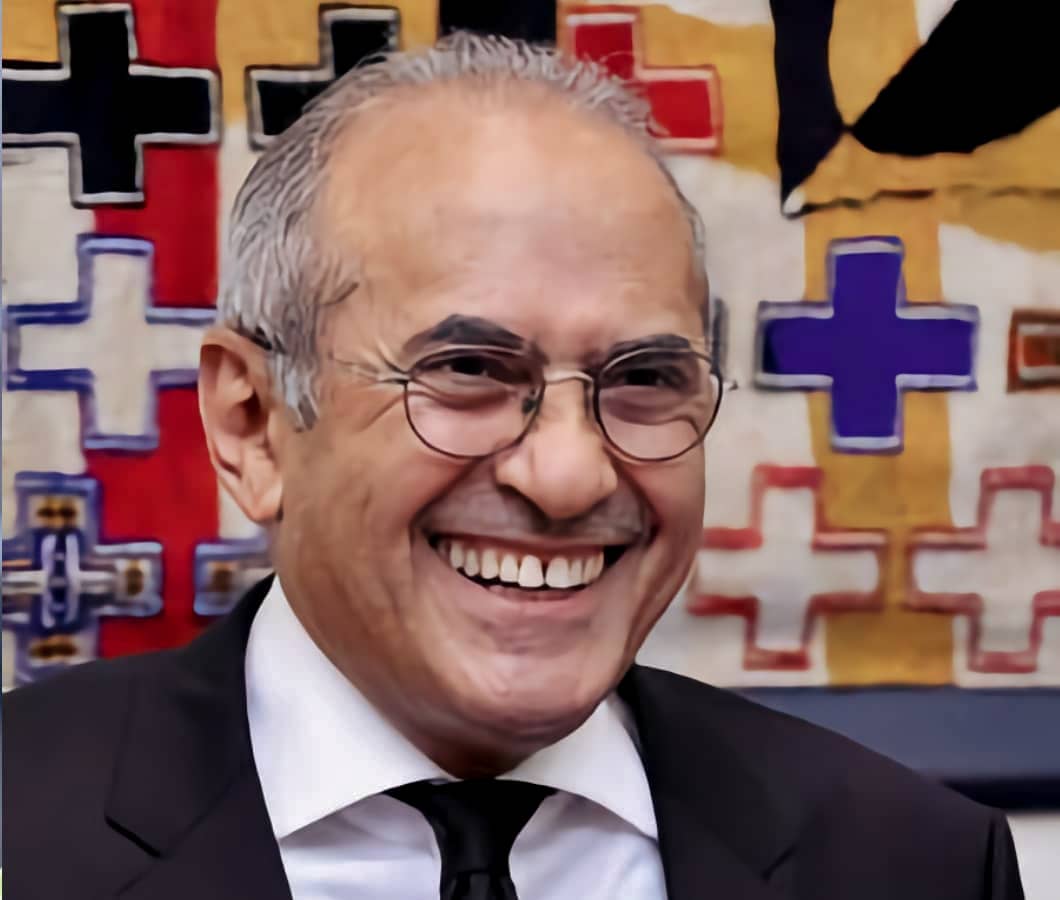Das Bild zeigt Scheich Mohammed Mussallam, einen lächelnden Mann mit Brille und grauen Haaren, der einen dunklen Anzug und eine Krawatte trägt, vor einem bunten Hintergrund mit Kreuzen in verschiedenen Farben.